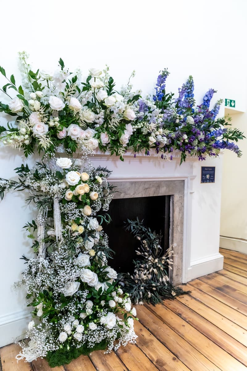 A white marble fireplace dressed by Hana Blossom in white roses, gypsophila and blue delphinium, flowers cascading down one side to the floor